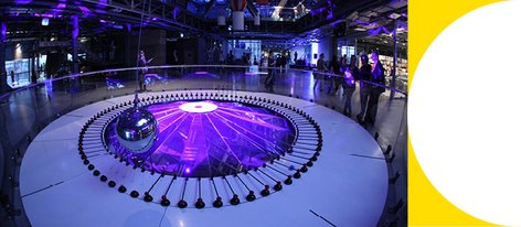  Wahadło Foucaulta w Centrum Nauki Kopernik otoczone fioletowym światłem, z widzami obserwującymi eksperyment.