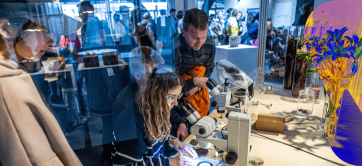  Grupa osób ogląda eksponaty i korzysta z mikroskopów przy stole w przestrzeni wystawowej; widoczne odbicia w szybie i kwiaty.