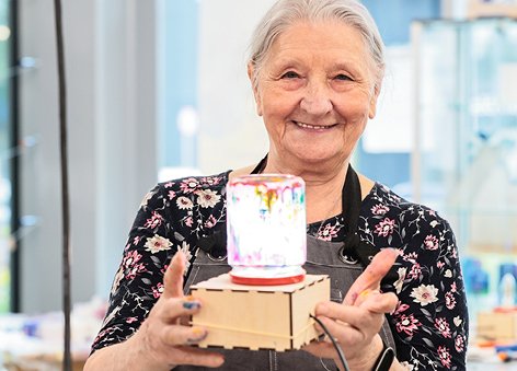  Starsza kobieta z uśmiechem prezentuje własnoręcznie wykonaną kolorową lampkę na drewnianej podstawie.