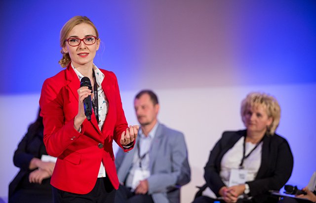 Na zdjęciu widać ubraną w czerwony żakiet Magdalenę Osial, która przemawia w czasie spotkania dla uczestników projektu. W tle inni występujący w czasie spotkania.