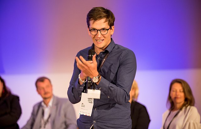 Na zdjęciu widać Dariusza Aksamita, ubranego w niebieską koszulę. Naukowiec przemawia, gestykulując, w czasie spotkania dla uczestników projektu. W tle widać rozmytych pozostałych występujących w czasie spotkania.