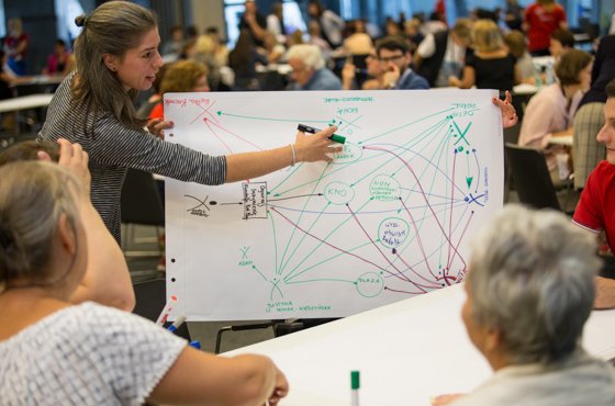 W tle wielu ludzi Na pierwszym planie kobieta rysuje na tablicy jakiś wykres.