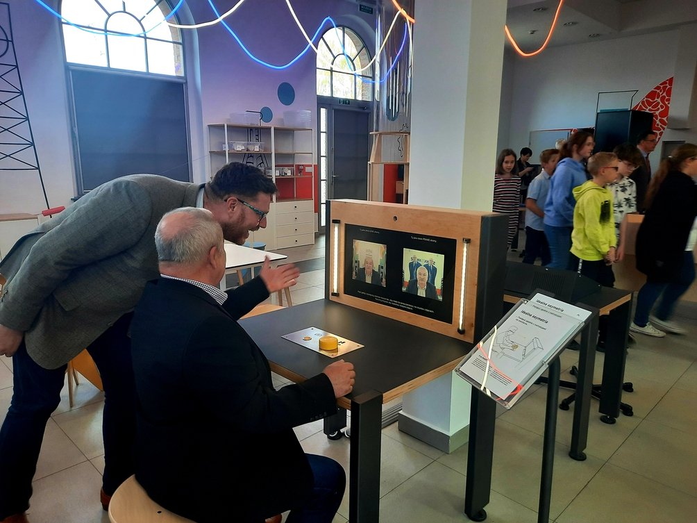 Dwóch mężczyzn przy eksponacie, gna którym widać zdjęcie twarzy jednego z nich.