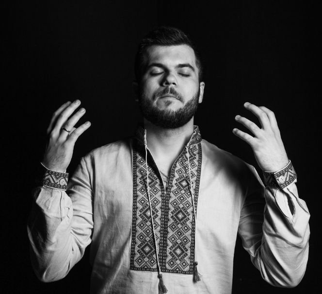 Czarno-białe zdjęcie artysty w tradycyjnej haftowanej koszuli ukraińskiej, ze wzniesionymi dłońmi.  