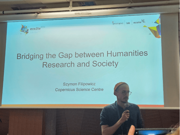 Szymon Filipowicz prezentujący slajd na konferencji. Slajd zatytułowany „Briding the Gap between Humanities Research and Society”.