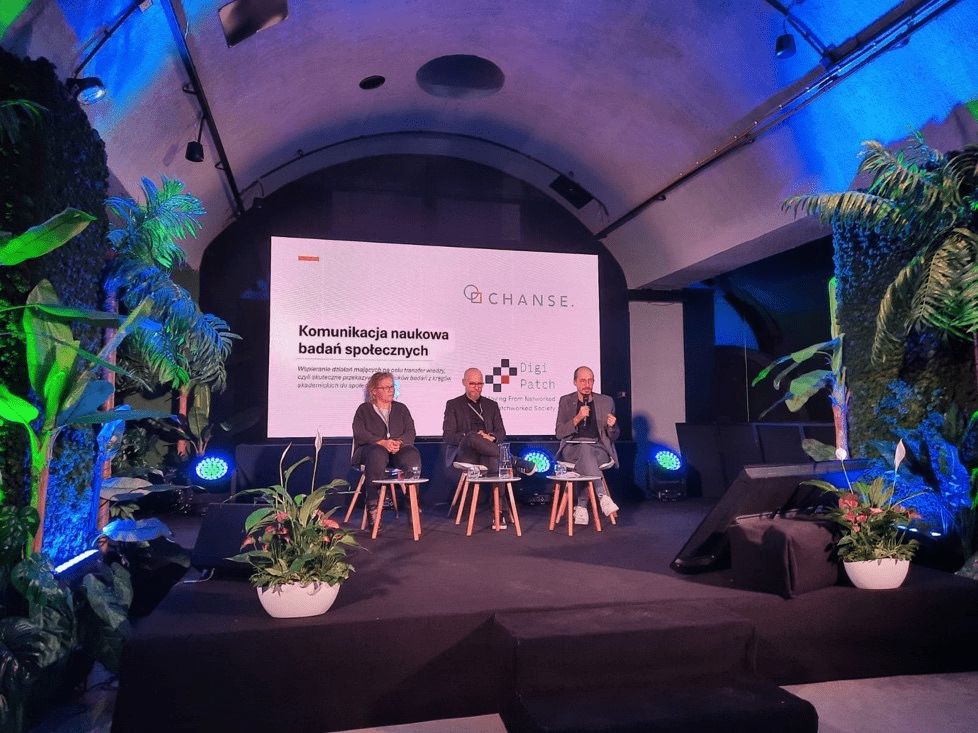 Zdjęcie przedstawia panel dyskusyjny w klimatycznej sali z roślinnymi dekoracjami. Na scenie znajduje się trzech prelegentów siedzących na drewnianych krzesłach, za nimi widoczny jest ekran z logotypem programu CHANSE oraz tytułem prezentacji 'Komunikacja naukowa badań społecznych'. W tle dominują zielone rośliny oraz nastrojowe oświetlenie w odcieniach niebieskiego.