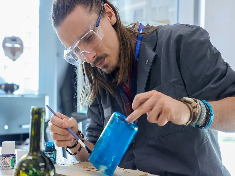  Mężczyzna w okularach ochronnych starannie maluje niebieski szklany słoik.
