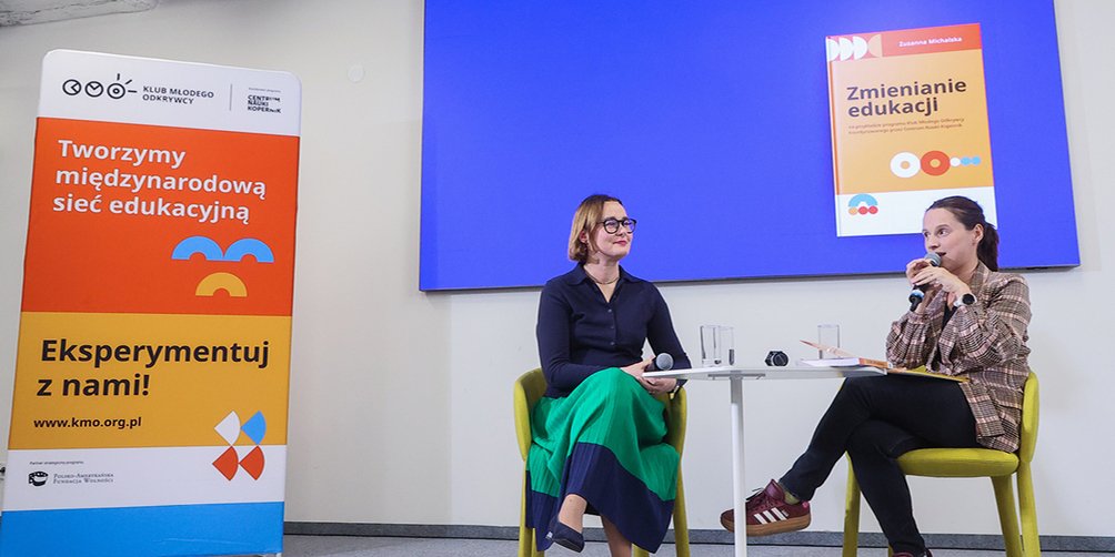 Dwie kobiety rozmawiają na scenie podczas spotkania o edukacji. Obok widoczny baner Klubu Młodego Odkrywcy i ekran z okładką publikacji „Zmienianie edukacji”.