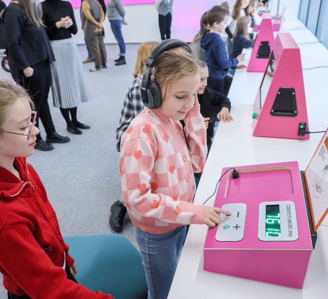 Dziewczynka w słuchawkach korzysta z różowego stanowiska, naciskając przyciski i obserwując wynik na wyświetlaczu, obok stoi koleżanka.