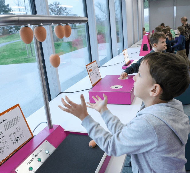 Chłopiec ćwiczy refleks, podrzucając zawieszone „jajka” na interaktywnym stanowisku przy oknie; w tle inni uczniowie wykonują doświadczenia.