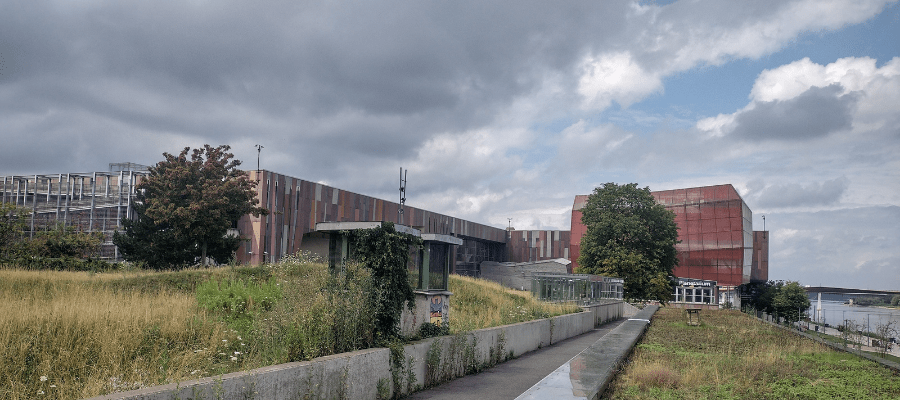 Widok na Centrum Nauki Kopernik i Planetarium od strony ogrodu. Na pierwszym planie ścieżka i dzika roślinność, w tle budynki i zachmurzone niebo.