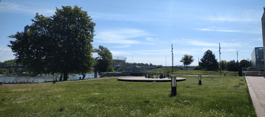 Zielony teren nad Wisłą obok Centrum Nauki Kopernik; na trawie okrągła platforma z roślinami, w tle drzewa, rzeka i stadion PGE Narodowy.