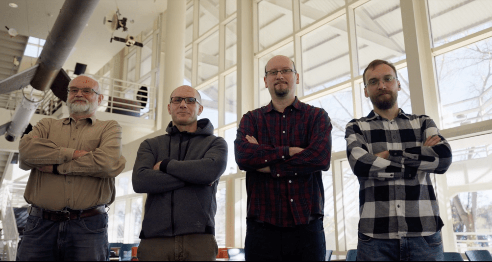 Czterech mężczyzn stoi z założonymi rękami w jasnym wnętrzu budynku; w tle duże okna i elementy ekspozycji naukowej, m.in. model sondy kosmicznej.