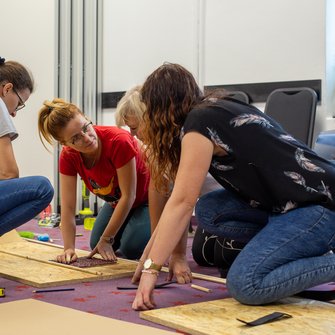 Na zdjęciu widać cztery kobiety pochylone nad podłogą i leżącymi na niej deskami i narzędziami.Kobiety rozmawiają podczas konstruowania. 