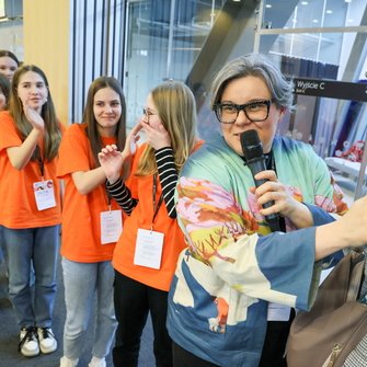 Grupa nastolatek w pomarańczowych koszulkach idzie za prowadzącą z mikrofonem