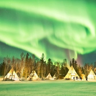 „Zielona zorza polarna tańczy nad oświetlonymi namiotami tipi w zimowym, leśnym krajobrazie.”