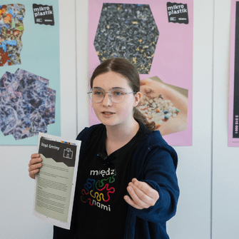 Uczennica prezentuje wyniki projektu o mikroplastiku, trzyma kartę pracy; w tle plansze edukacyjne i wykresy.