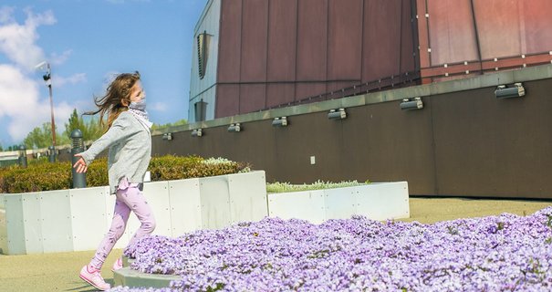 Biegnąca dziewczynka z szeroko rozłożonymi rękami, wokół niej fioletowe kwiaty.