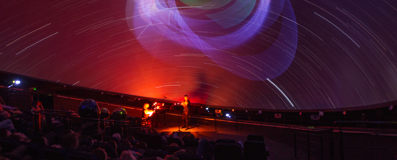 Widok sceny pod kopułą Planetarium, wszystko jest w czerwonym świetle, na scenie kobieta przy fortepianie i mężczyzna gra na skrzypcach.