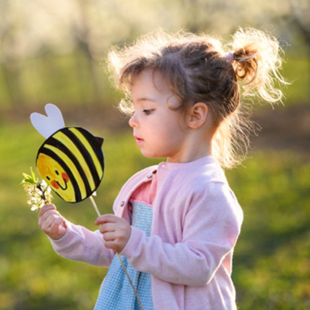 Zdjęcie dziewczynki w różowym sweterku, bokiem, na zielonym tle. Dziewczynka w ręce trzyma papierową pszczołę.