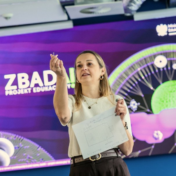 Nauczycielka trzyma diodę w ręce, pokazuje ją, w tle fioletowa i zielona kolorystyka  ZBADAI