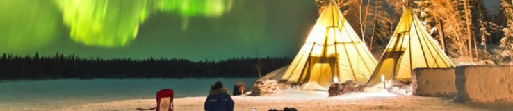  Zorza polarna tańcząca nad zimowym obozowiskiem z tipi i osobą siedzącą na śniegu.