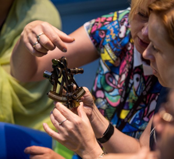 Na środku kadru jakiś eksponat, podobny o sekstansu. Trzymają go dwie kobiety.