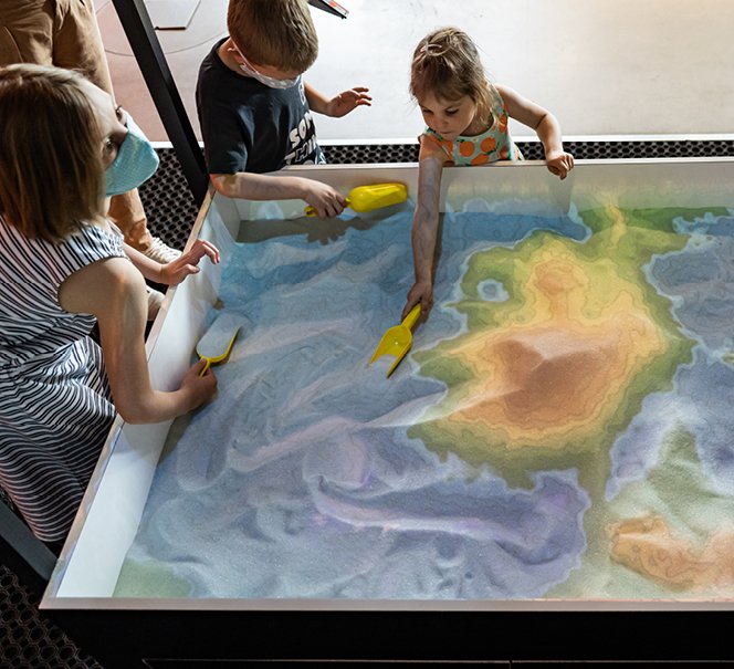 Ludzie przy eksponacie "Usyp ziemię" - piasek podświetlony na kolorowy przypomina mapę.