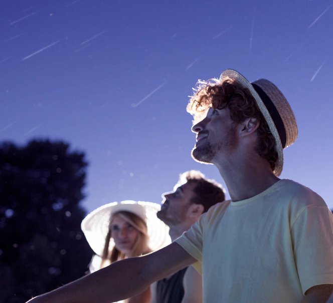 Troje ludzi w letnich strojach wpatrzonych w nocne niebo, na którym widać spadające gwiazdy.
