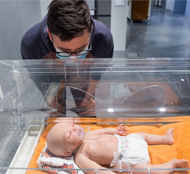 Człowiek pochyla się nad inkubatorem, w którym leży manekin przypominający niemowlę.