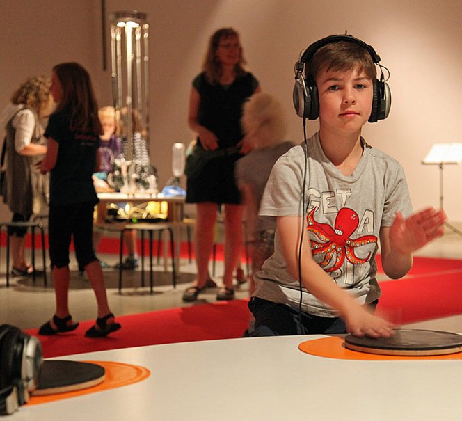 Chłopiec w słuchawkach miksujący płytę, z tyłu widać postaci innych zwiedzających.