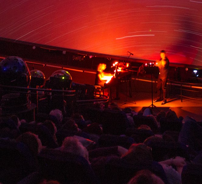 Widok sceny pod kopułą Planetarium, wszystko jest w czerwonym świetle, na scenie kobieta przy fortepianie i mężczyzna gra na skrzypcach.