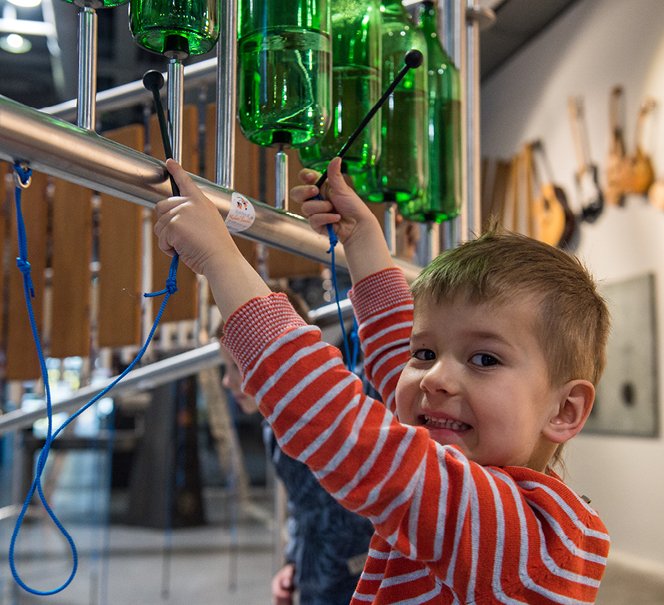Chłopiec odwracający twarz do fotografa, bawi się eksponatem muzycznym z zielonymi butelkami wypełnionymi wodą.