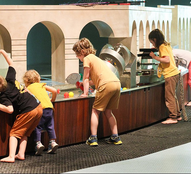 Grupa dzieci pochyla się nad długą instalacją wodną z kolorowymi piłeczkami.