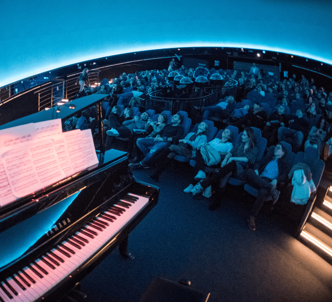 Widok z boku na publiczność w planetarium; na pierwszym planie fortepian z otwartą partyturą.