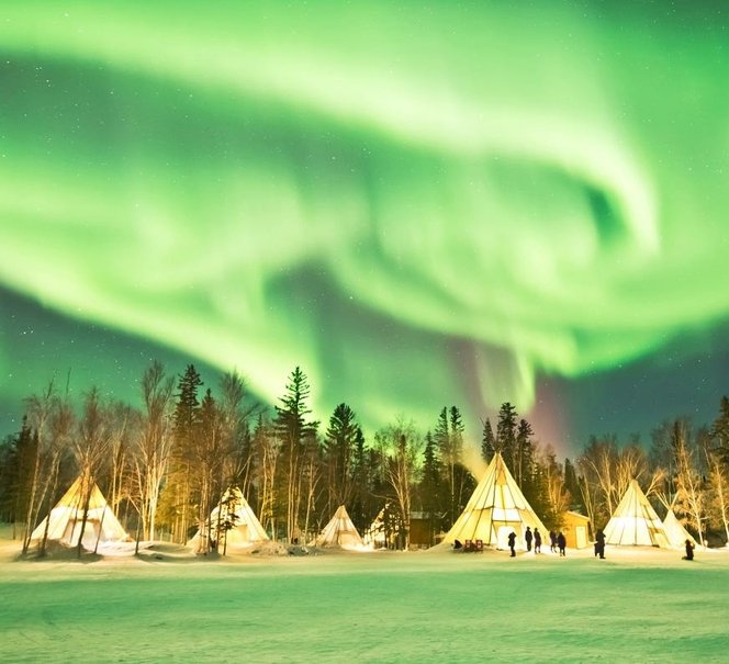 „Zielona zorza polarna tańczy nad oświetlonymi namiotami tipi w zimowym, leśnym krajobrazie.”