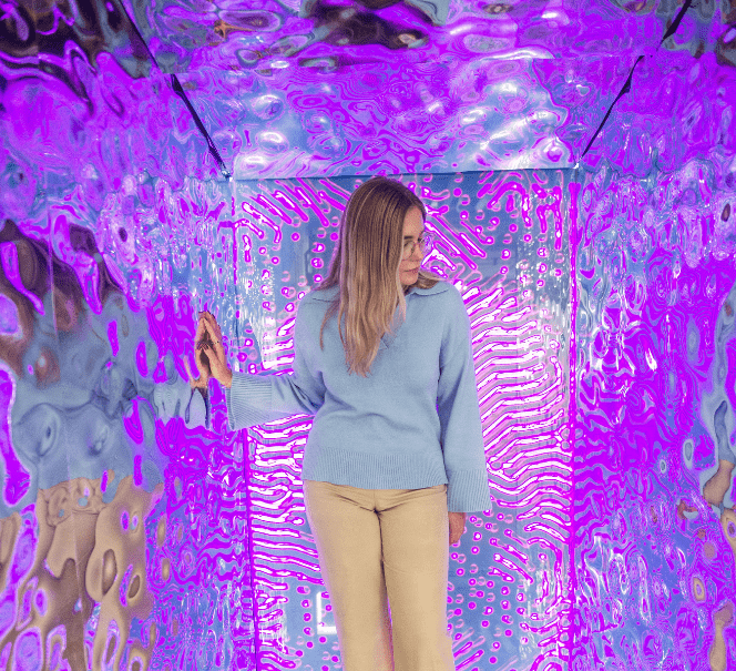 Kobieta w błękitnym swetrze stoi w świetlistej instalacji o fioletowych, falujących ścianach.