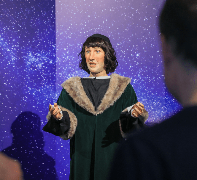 Postać w historycznym stroju z futrzanym kołnierzem stoi na tle rozgwieżdżonego nieba, prezentując gestami ideę lub odkrycie przed widownią.