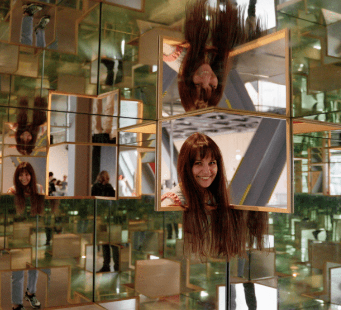Kobieta w instalacji z lustrami – jej odbicie powiela się w wielu kierunkach, tworząc efekt kalejdoskopu.