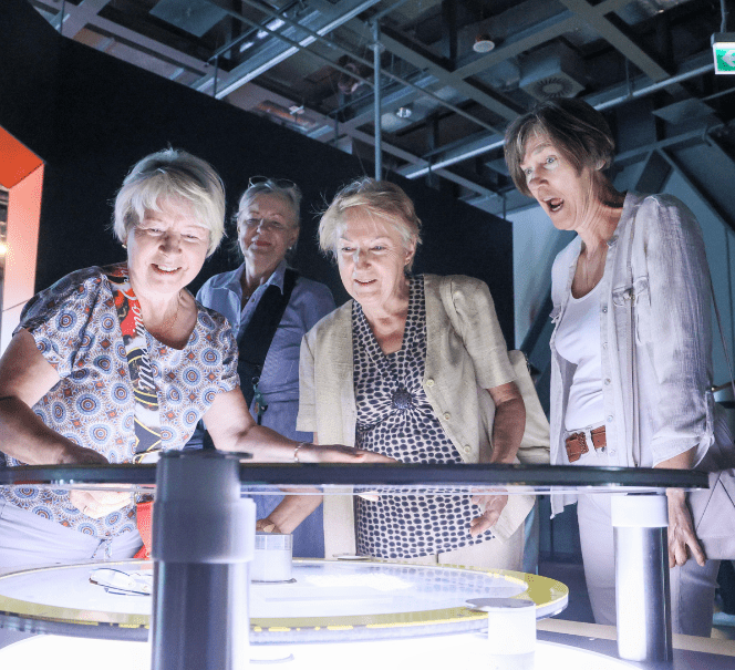 Grupa starszych kobiet z zainteresowaniem obserwuje świetlny eksponat w centrum nauki.