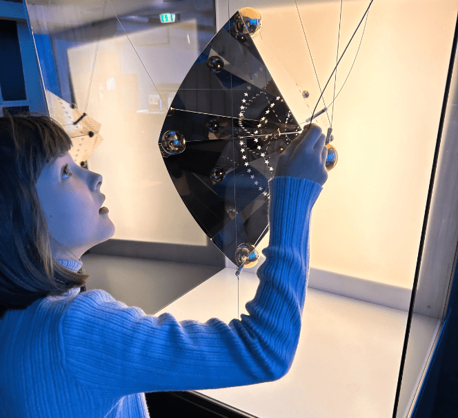 Dziecko dotyka instalacji z czarną powierzchnią i złotymi punktami połączonymi cienkimi nićmi.