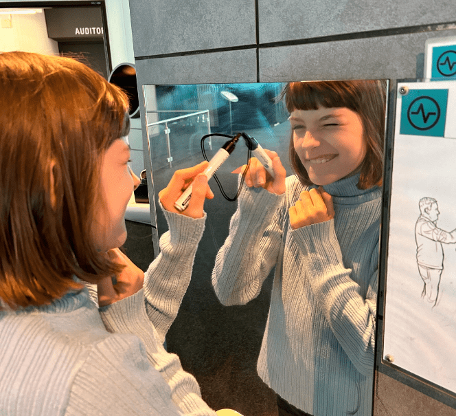 Dziecko rysuje po lustrze markerem, uśmiechając się do swojego odbicia.