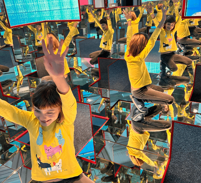 Dziecko w żółtej koszulce siedzi w przestrzeni pełnej luster, które zwielokrotniają jego postać.