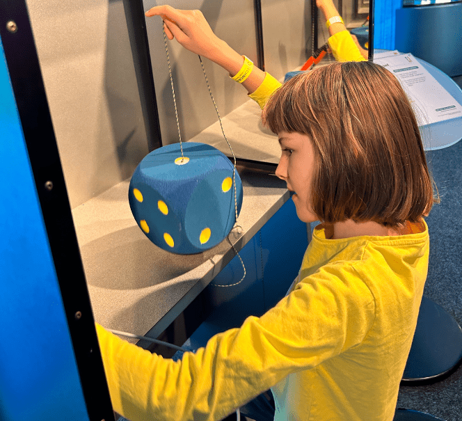 Dziecko w żółtej koszulce bawi się dużą zawieszoną kostką z niebieskimi i żółtymi ściankami.