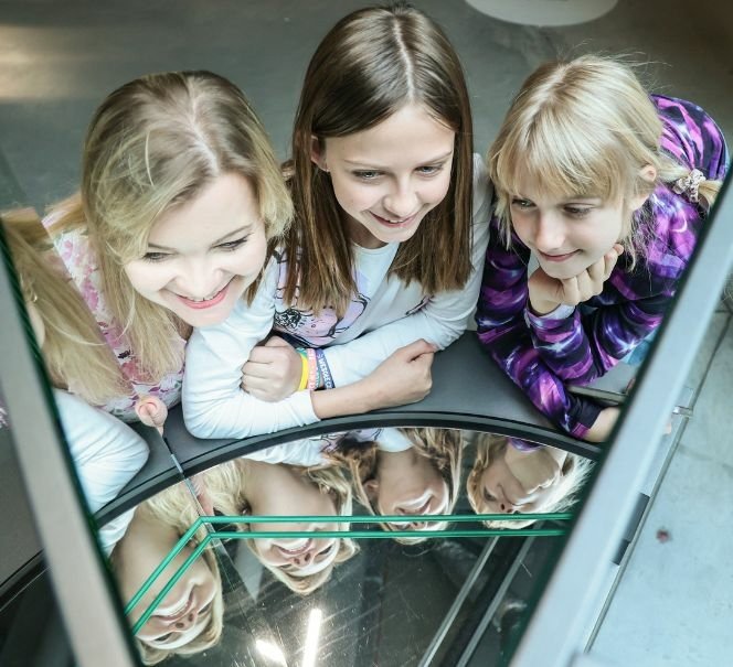 Trzy dziewczynki zaglądają do urządzenia z lustrami, w którym odbija się ich twarze.