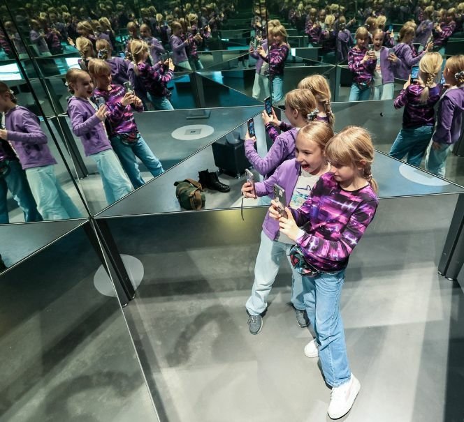 Dwie dziewczynki robią sobie zdjęcie w pomieszczeniu pełnym luster, które tworzą liczne odbicia.