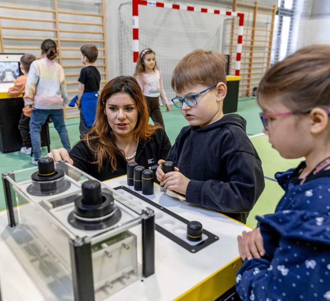 dukatorka tłumaczy dwójce dzieci działanie interaktywnego stanowiska naukowego w sali gimnastycznej; w tle inni uczniowie biorą udział w zajęciach.