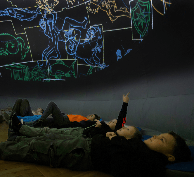 Dzieci leżące na podłodze w ciemnym pomieszczeniu, pokazują rękami kolorowe gwiazdozbiory nad głowami.