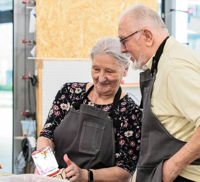 Starsza kobieta i starszy mężczyzna w fartuchach warsztatowych oglądają kolorowy przedmiot wykonany podczas zajęć w pracowni.