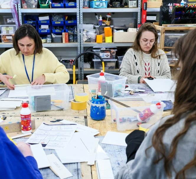 Dwie kobiety siedzą przy stole warsztatowym i pracują z papierem i narzędziami plastycznymi; wokół pojemniki z materiałami.